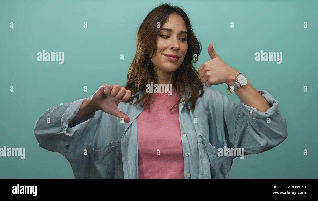 Woman expressing mixed emotions with thumbs up and down against a vibrant green background, showcasing diverse communication and mood concept. Stock Photo
