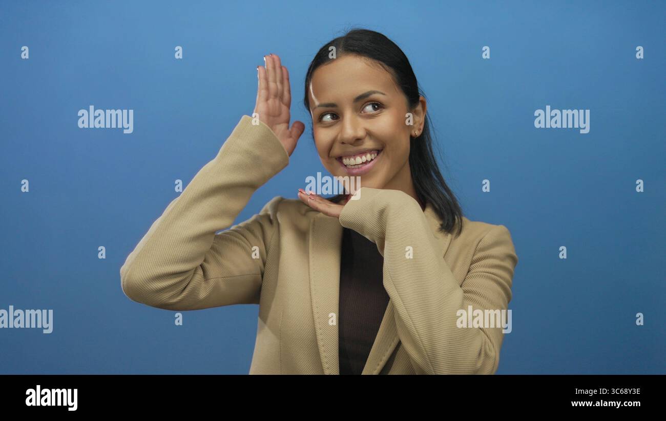 Woman smiling against blue background with hands framing face wearing beige jacket representing happiness and youthful energy in an isolated studio se Stock Photo