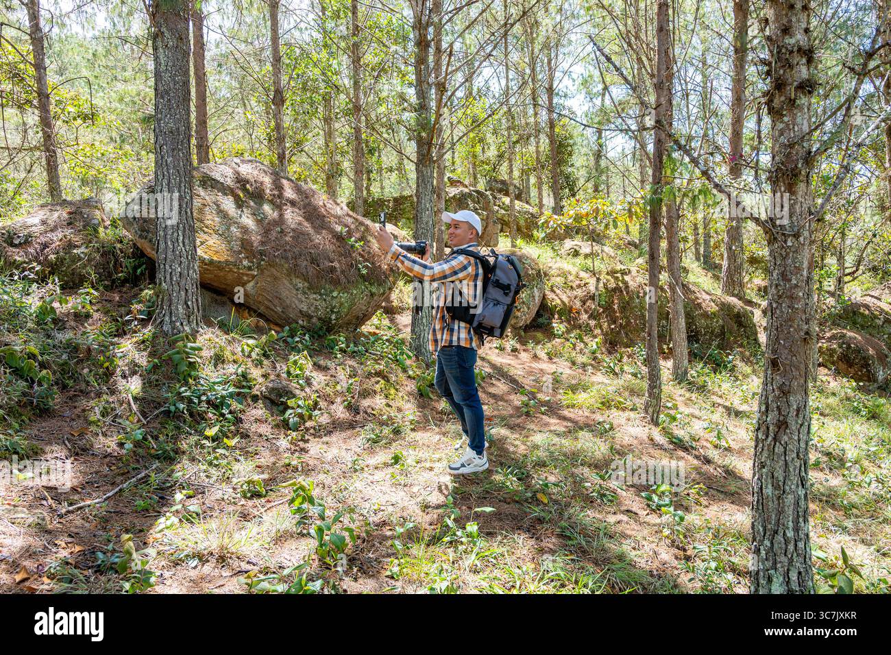 Photographer in a pine forest smiling as he takes a selfie with one hand and holds his camera in the other, capturing the behind-the-scenes of his adv Stock Photo