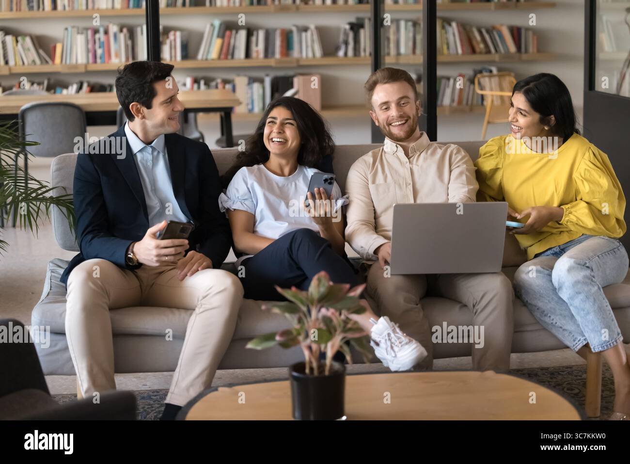 Four colleagues holding diverse devices talking seated on sofa Stock Photo