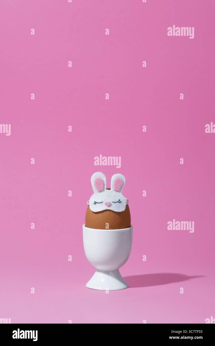 Brown egg in bunny sleep mask in a white egg cup on pink background. Easter banner, place for your text. Stock Photo