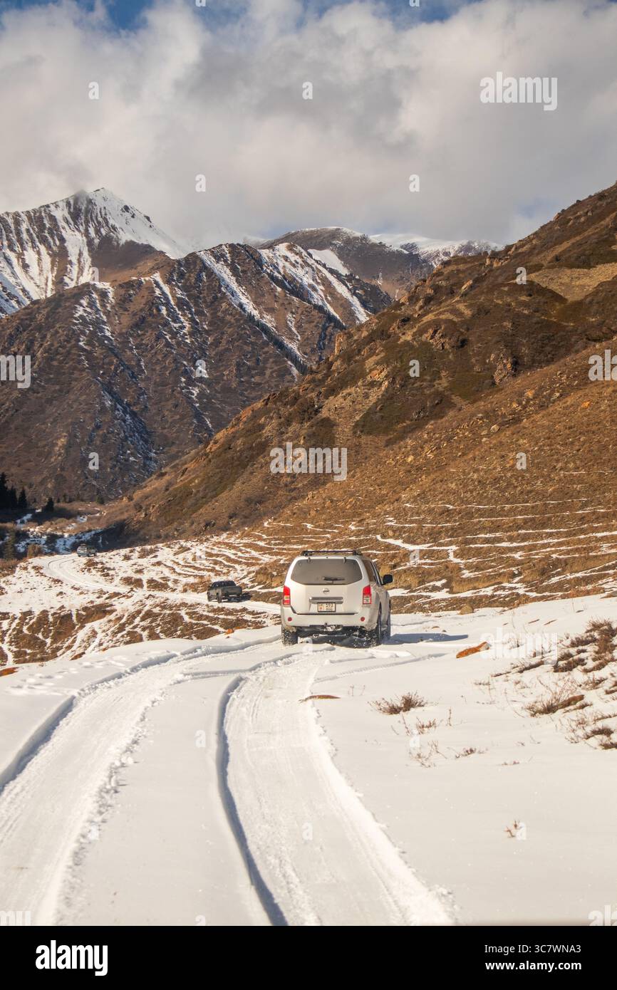 Naryn, Kyrgyzstan - October 12, 2024: Two off-road vehicles drive along a snowy mountain road in a vast valley with a snowcapped mountain range in the Stock Photo