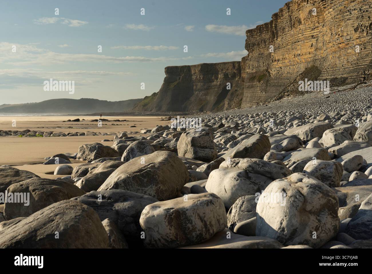 A lone person explores the vast sandy beach at low tide, backed by the imposing cliffs of Monknash Beach, Wales. Stock Photo