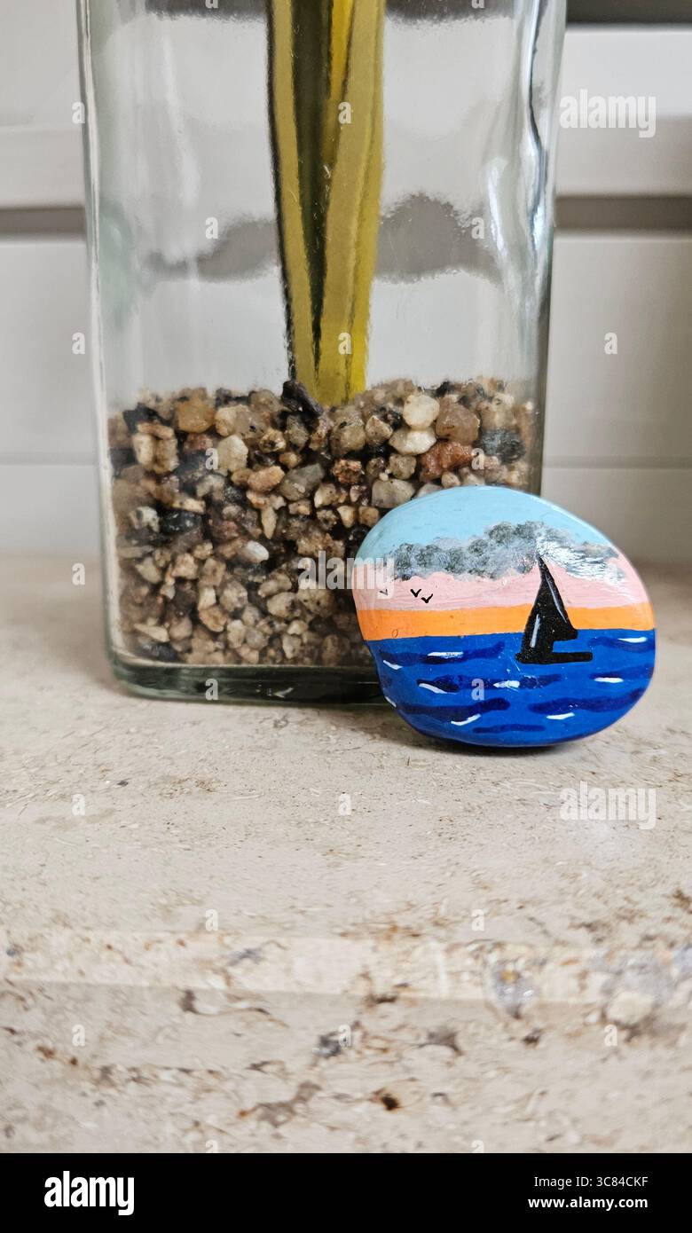 A hand-painted stone showcasing a serene seascape with a small sailboat floating on calm blue waters, framed by a glowing sunset sky and flying birds. - Smartphone Captured Stock Image