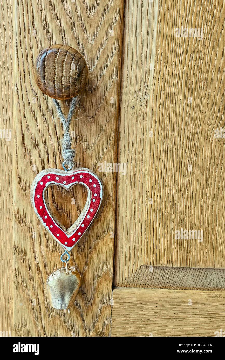 A red and white polka-dotted heart-shaped wooden ornament with a small bell hangs from a rope on a wooden cabinet handle. - Smartphone Captured Stock Image