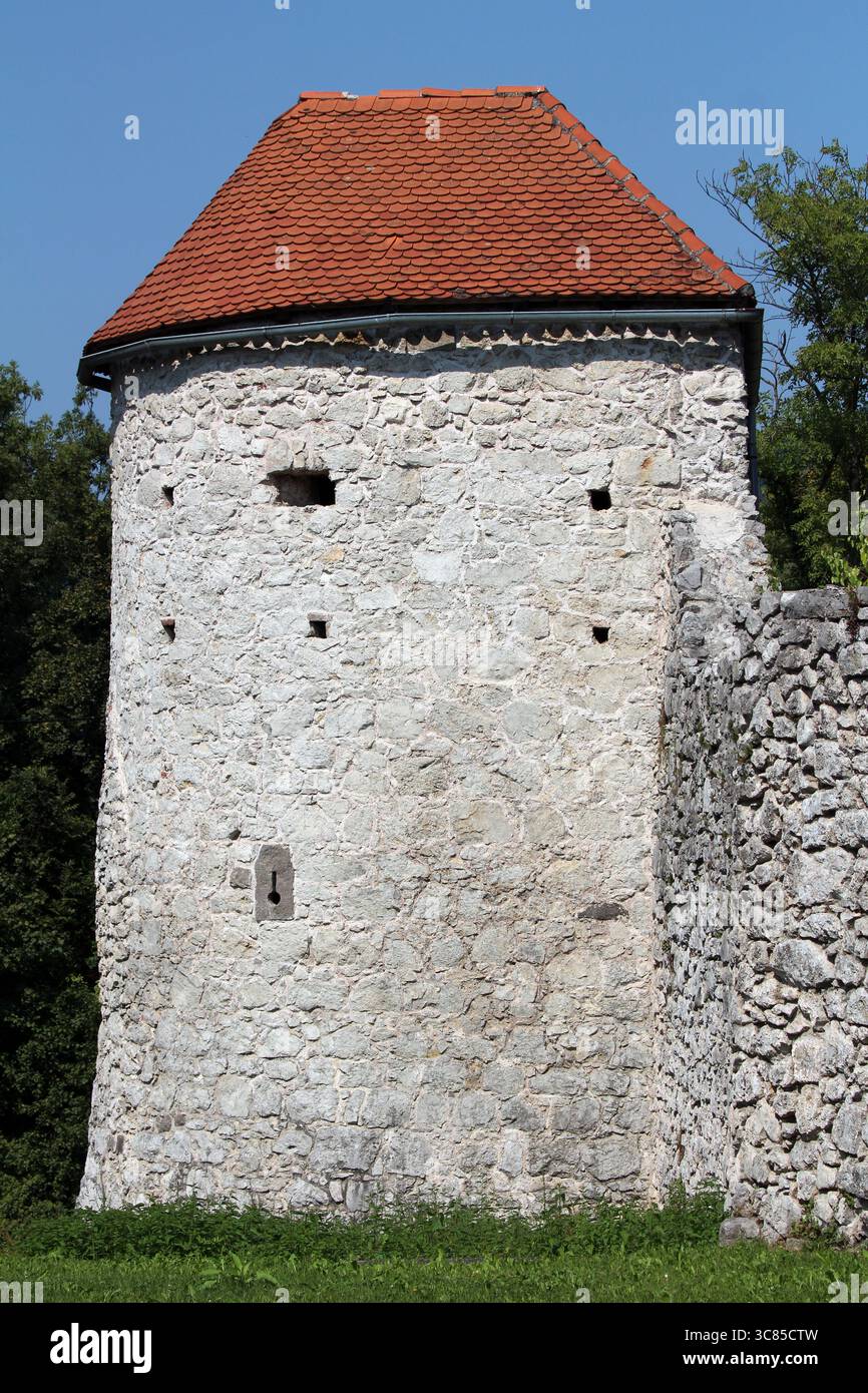 Ancient watchtower built of rough stone with red tile roof, small slit windows for archers, and attached stone wall, surrounded by greenery Stock Photo
