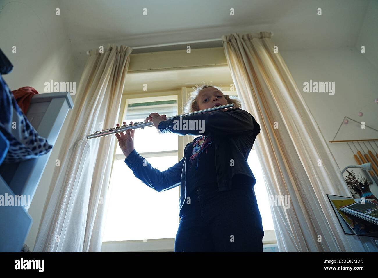 Young musician passionately plays the flute in a bright room with sunlight streaming through the window. Wearing casual outfit, they focus intently on Stock Photo