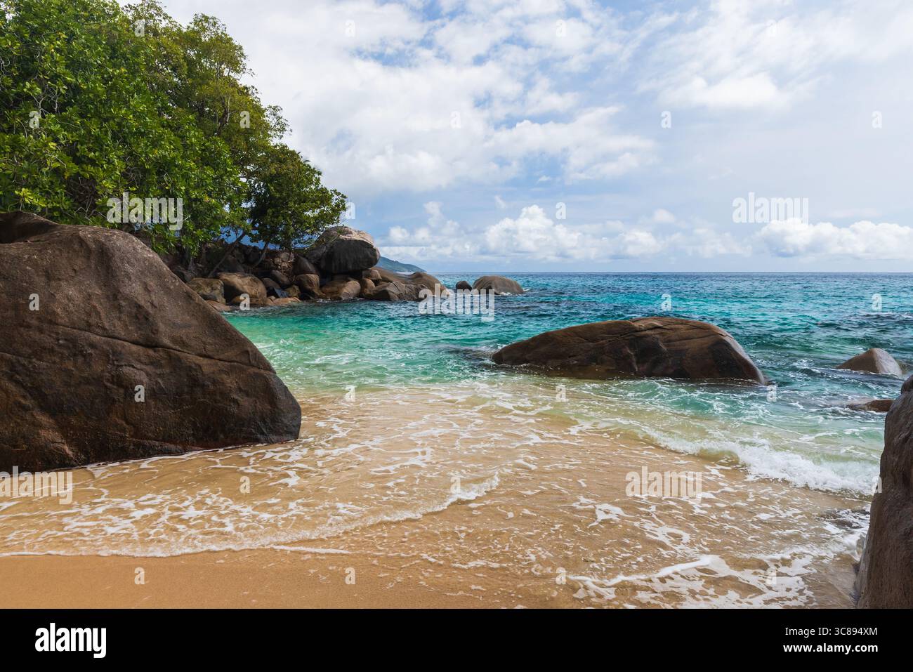 Mahe island, Seychelles. A tranquil tropical beach scene featuring pristine golden sand, clear turquoise waters, large boulders, and lush greenery und Stock Photo