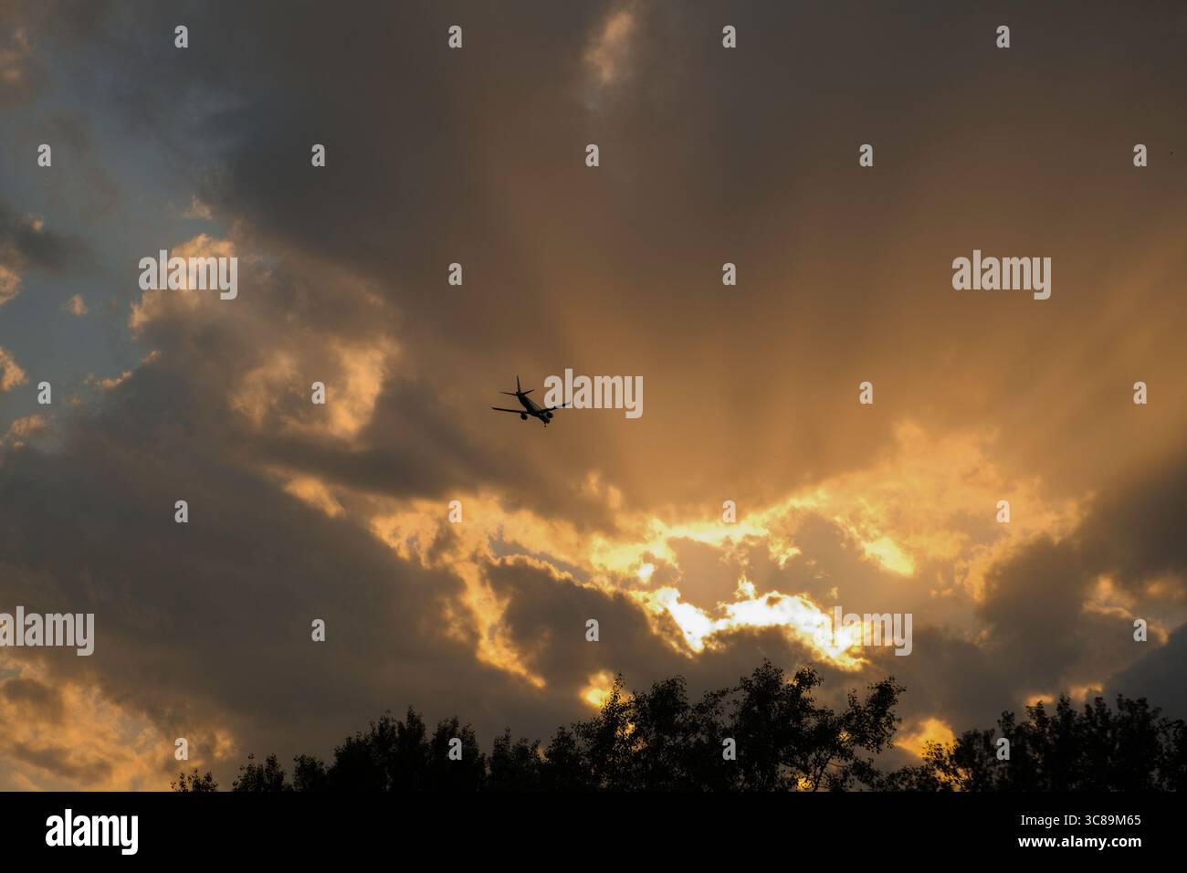 Airplane flying through dramatic sunset clouds with sun rays breaking through Stock Photo