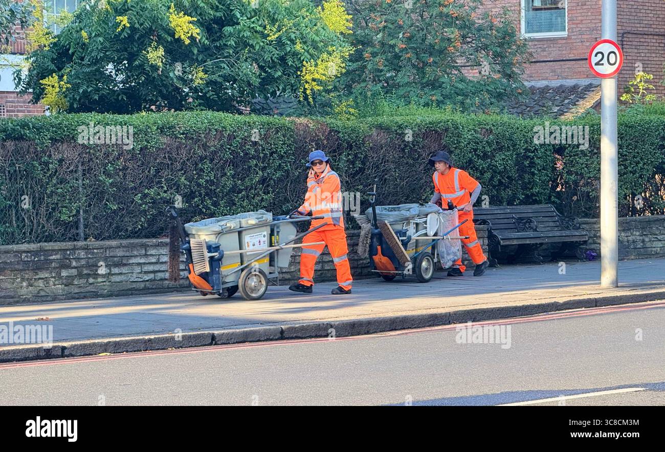 Two London street cleaners dressed in orange reflective clothing, walk up a road pushing carts containing their cleaning gear. - Smartphone Captured Stock Image