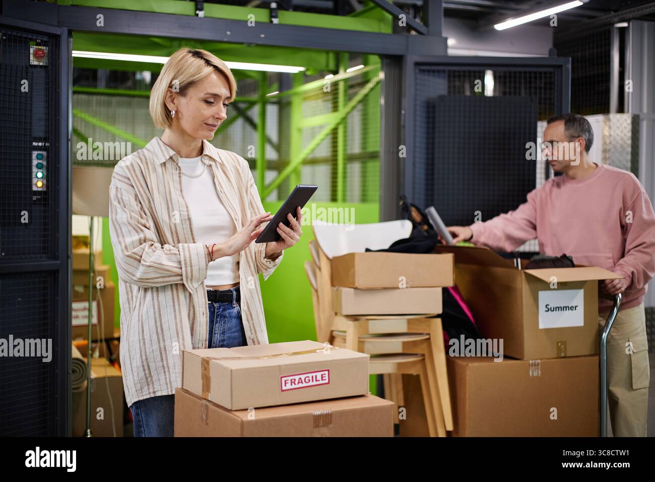 Caucasian Middle Aged Woman Using Digital Tablet While Packing Cardboard Boxes Stock Photo