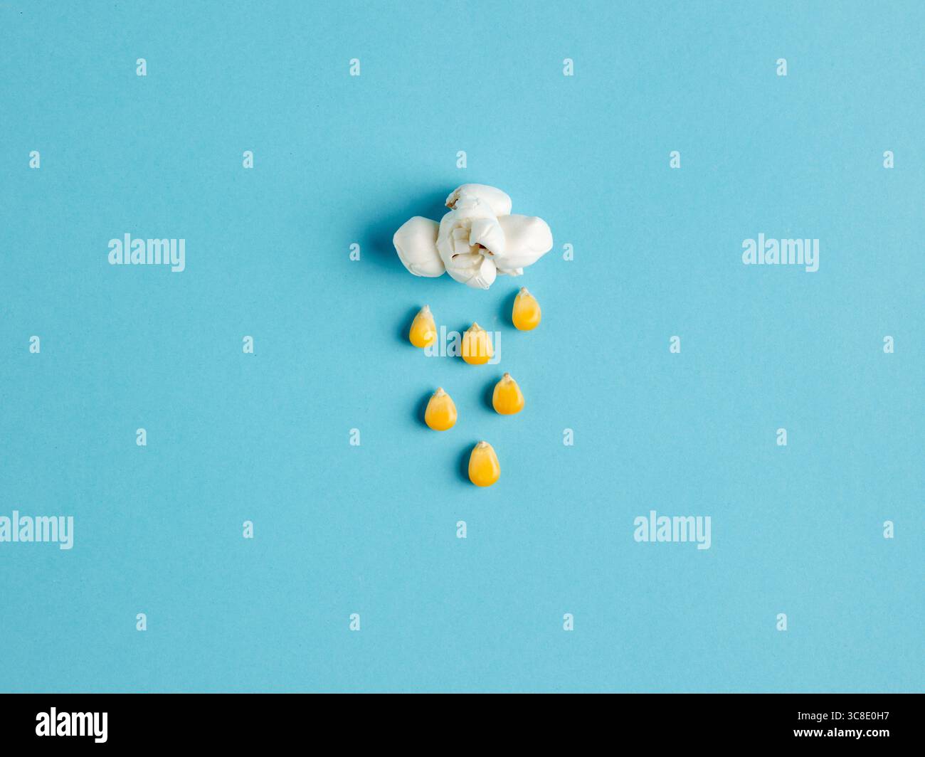Creative arrangement of popcorn and corn kernels shaped like a cloud raining corn over a vibrant blue background, symbolizing rain clouds. Stock Photo
