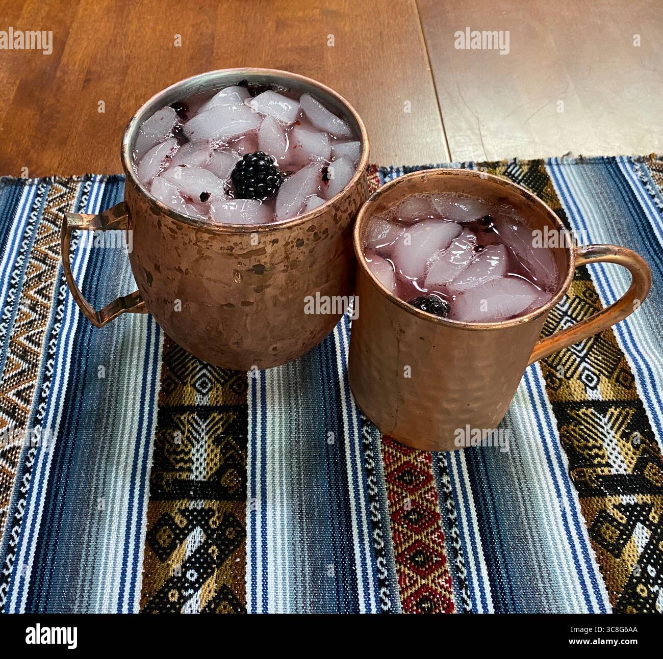 Two copper mugs, filled with ice, blackberries and vodka, sitting on a hand woven placemat, on a wood table - Smartphone Captured Stock Image
