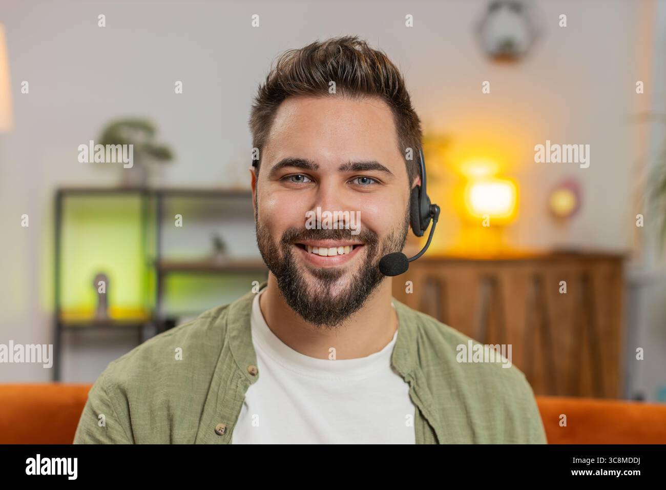 Confident young Caucasian man sitting on sofa at home wearing headset, freelance worker, call center, support service operator helpline, having talk with client or colleague, communication support. Stock Photo