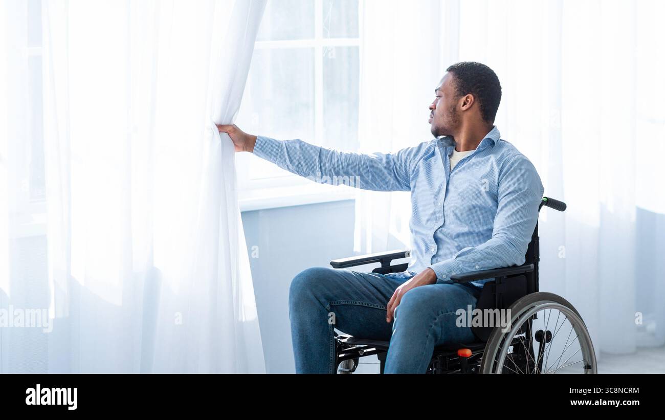 Sad with disability young man sitting on wheelchair alone at home, looking through window Stock Photo