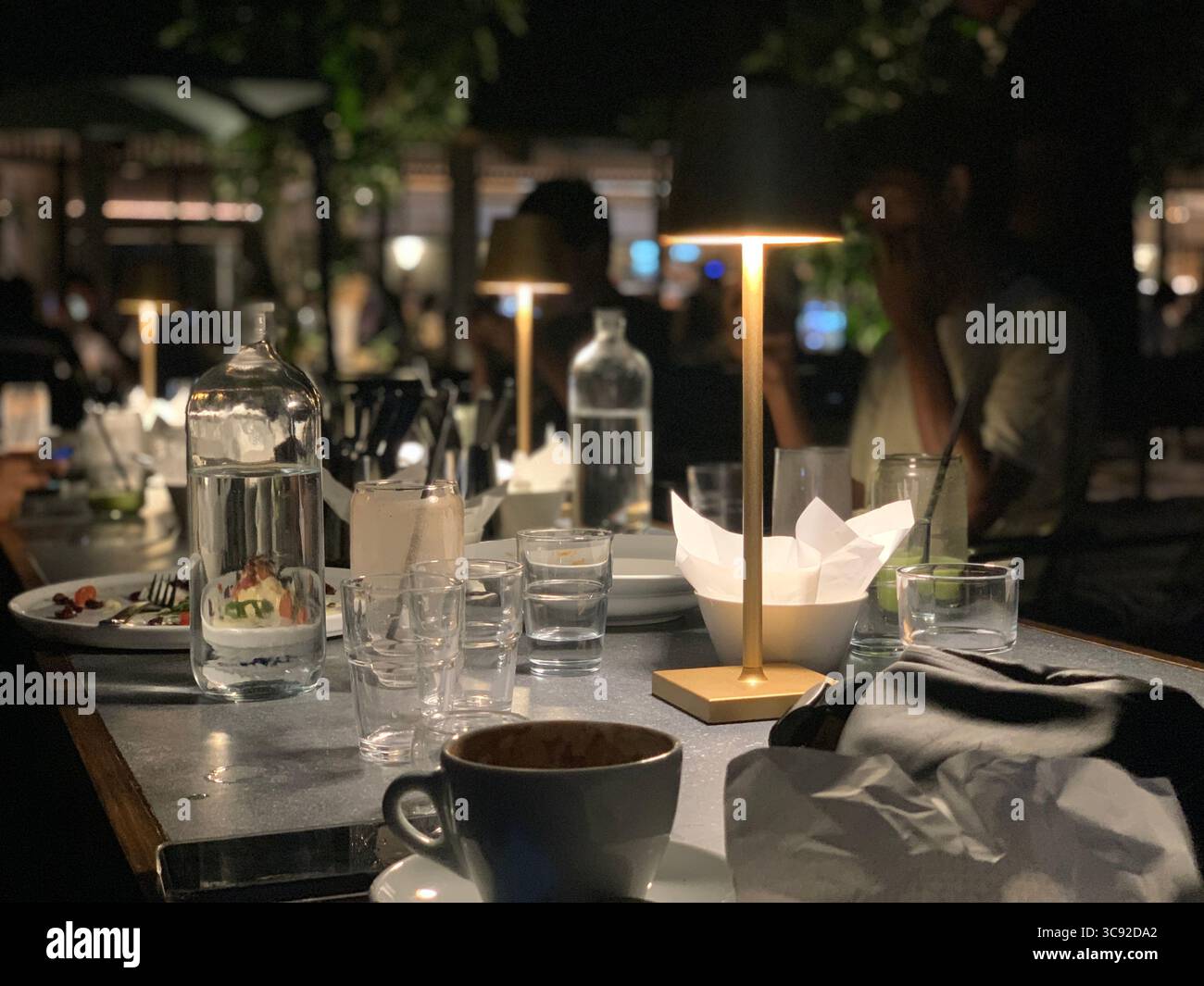 Late-night restaurant table scattered with cups, bottles and warm lamp light; diners blur into the background — an intimate snapshot of evening dining - Smartphone Captured Stock Image