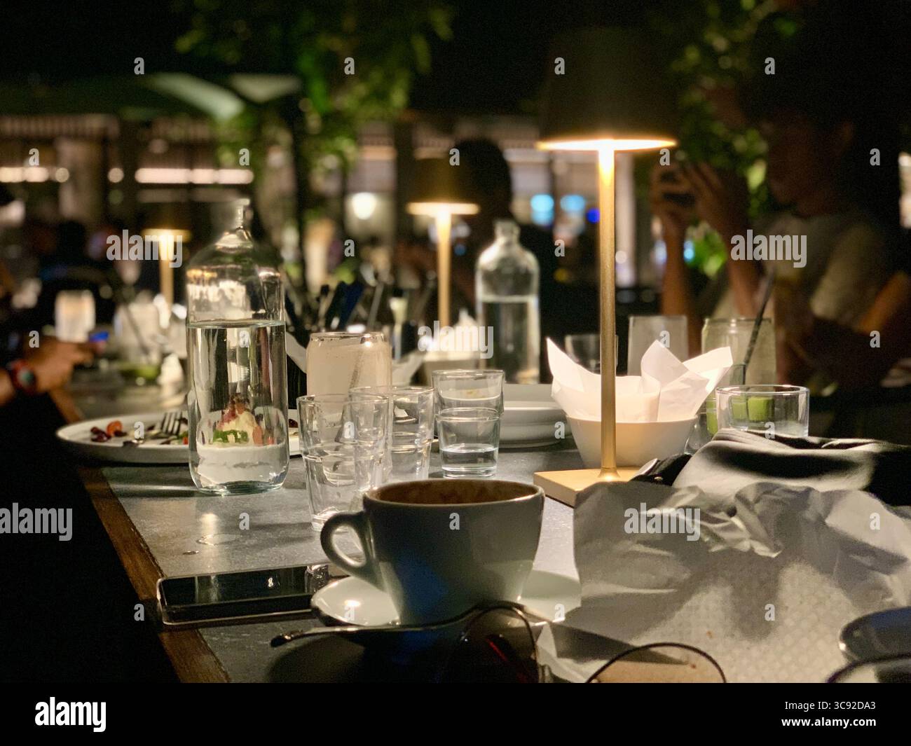 Evening dining scene: a restaurant table with cups, bottles and soft lamps; diners in the background create a lively, intimate night atmosphere. - Smartphone Captured Stock Image