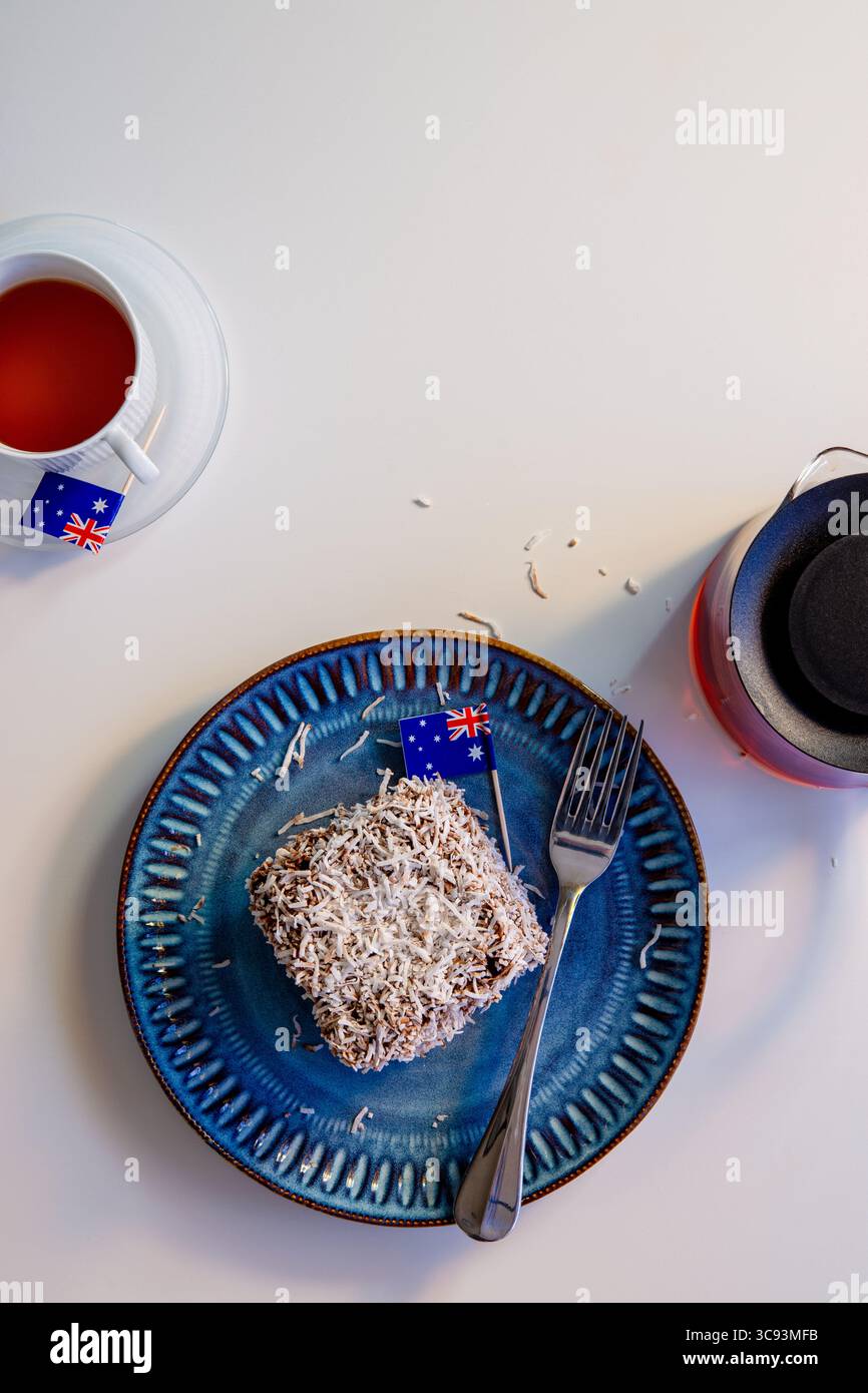 A Lamington cake with two Australian flags, one on the cake and one on a teacup saucer. A fork, blue plate, and glass teapot are on the white table. Stock Photo