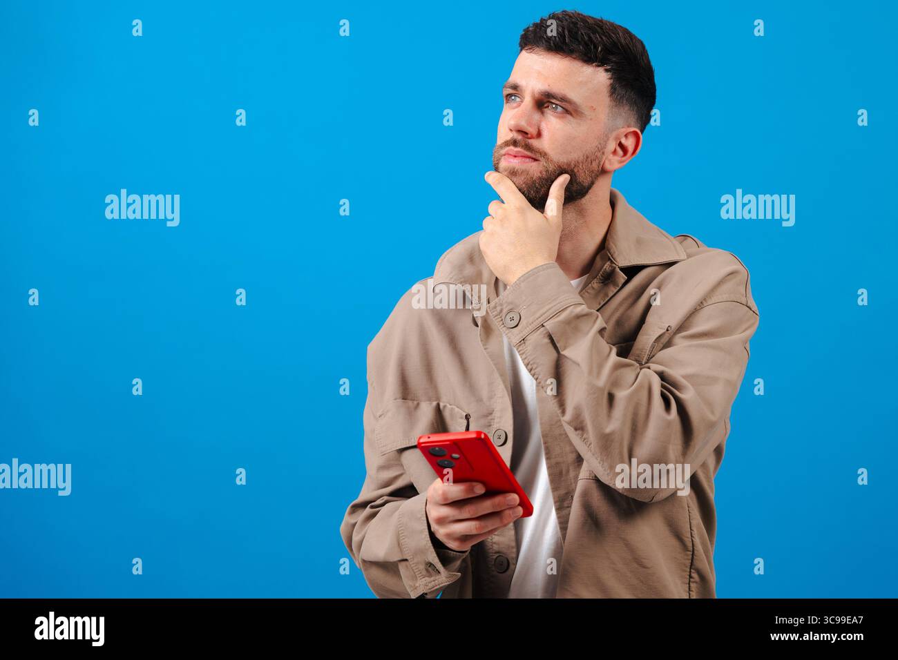 Pensive young man with smartphone over blue backdrop, planning next message idea Stock Photo