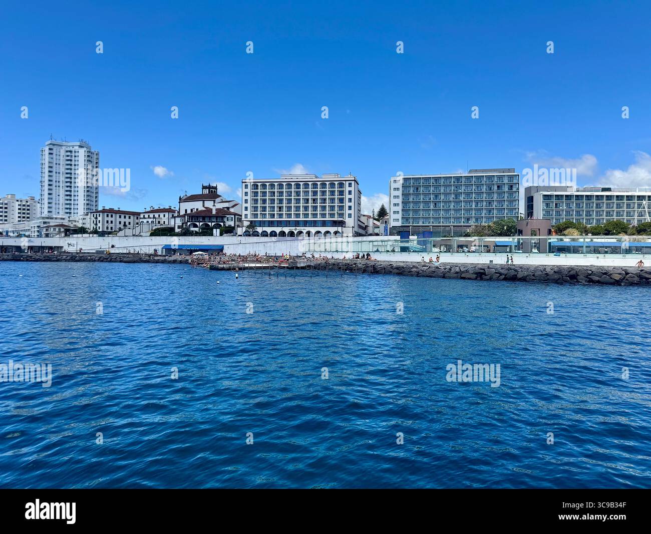 Seafront view of Ponta Delgada city, São Miguel Island, Azores, Portugal, featuring modern buildings, a church, and the popular ocean swimming area on - Smartphone Captured Stock Image