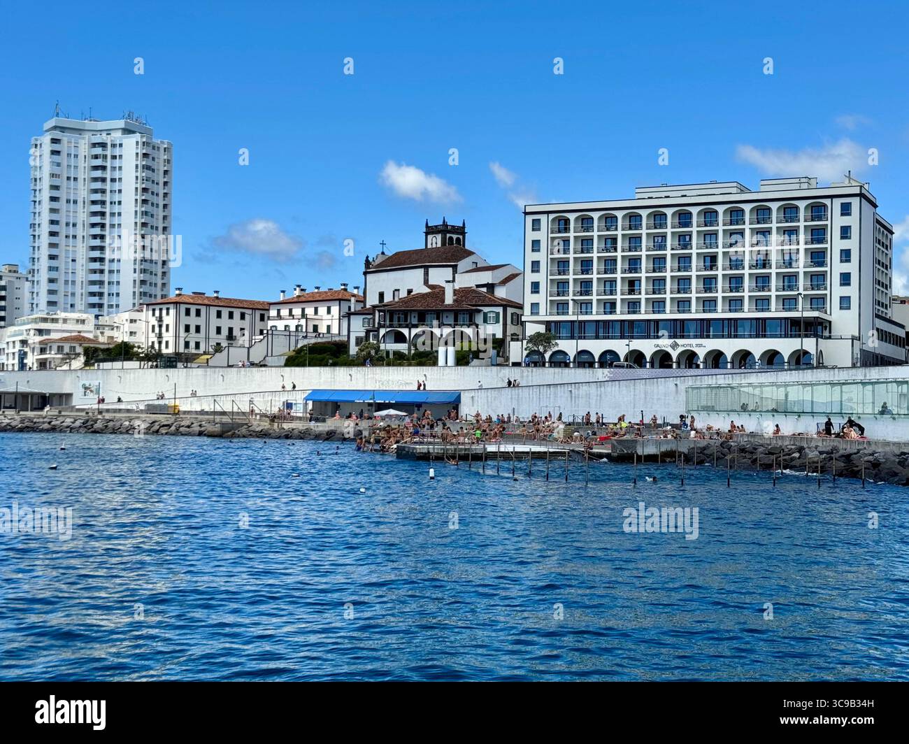 Seafront view of Ponta Delgada city, São Miguel Island, Azores, Portugal, featuring modern buildings, a church, and the popular ocean swimming area on - Smartphone Captured Stock Image