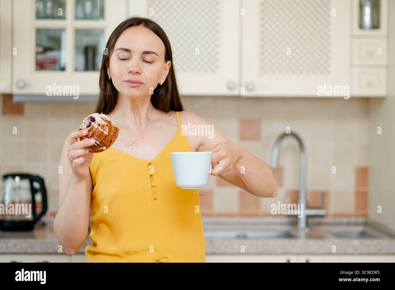Peaceful moment as a young woman enjoys tea and a sweet bun in a warm, cozy kitchen, symbolizing comfort and slow living Stock Photo