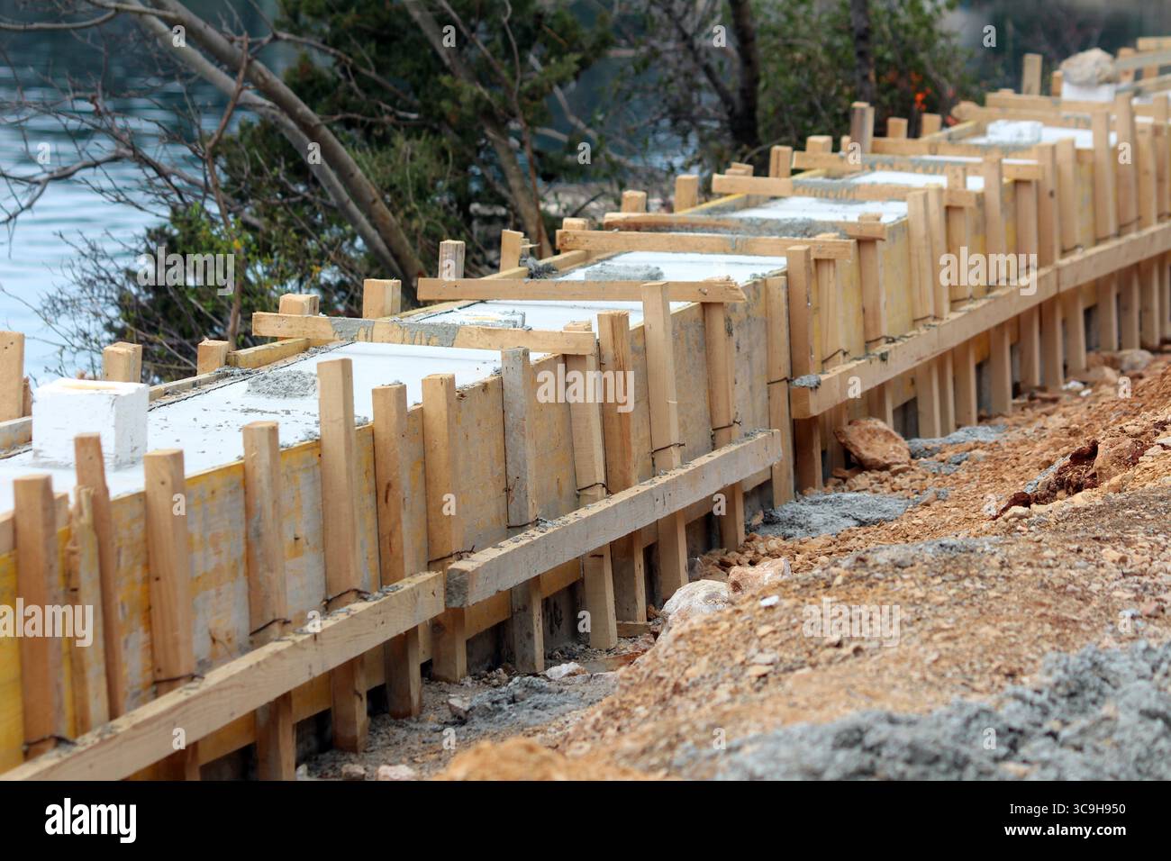 New concrete is setting inside sturdy wooden formwork, creating a straight foundation line for a riverside path, surrounded by soil and sparse Stock Photo