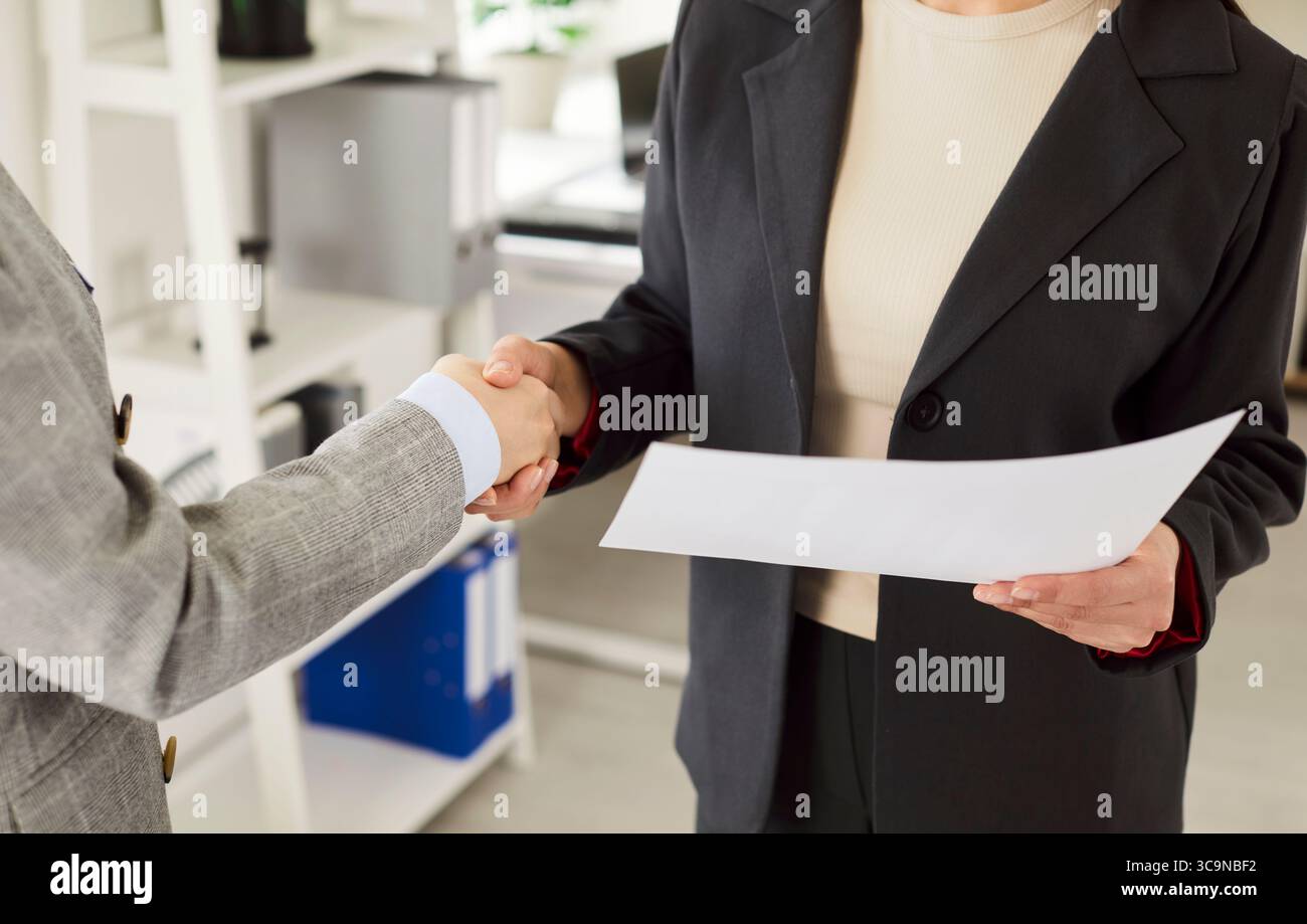 Two businesswomen in formal suits holding contract, making agreement and shaking hands in office. Stock Photo
