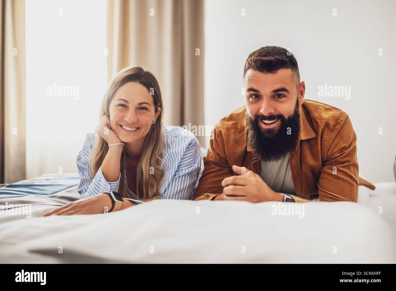 Young happy couple is lying on bed in their bedroom, looking at camera and smiling. Stock Photo