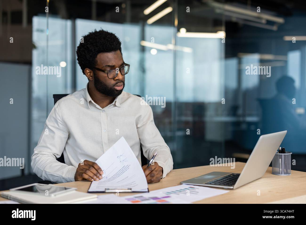 Young professional man engaged in reviewing documents while seated at a desk in a modern office environment. He displays focus and dedication, utilizing a laptop and papers to complete his tasks. Stock Photo