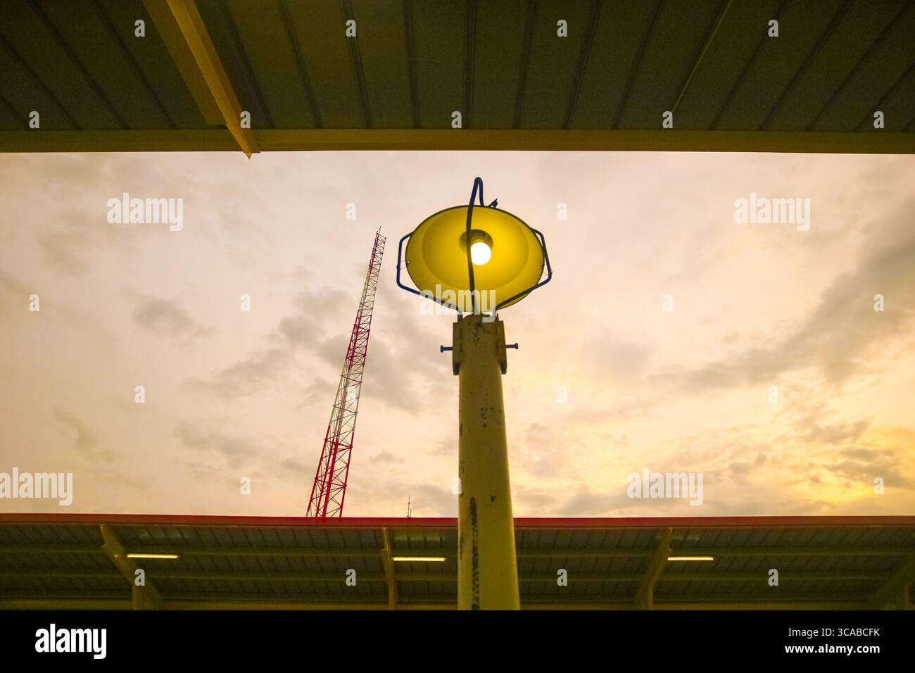 Street lamp with warm glow beside red and white radio tower under golden sunset sky, merging industry with evening beauty. Stock Photo