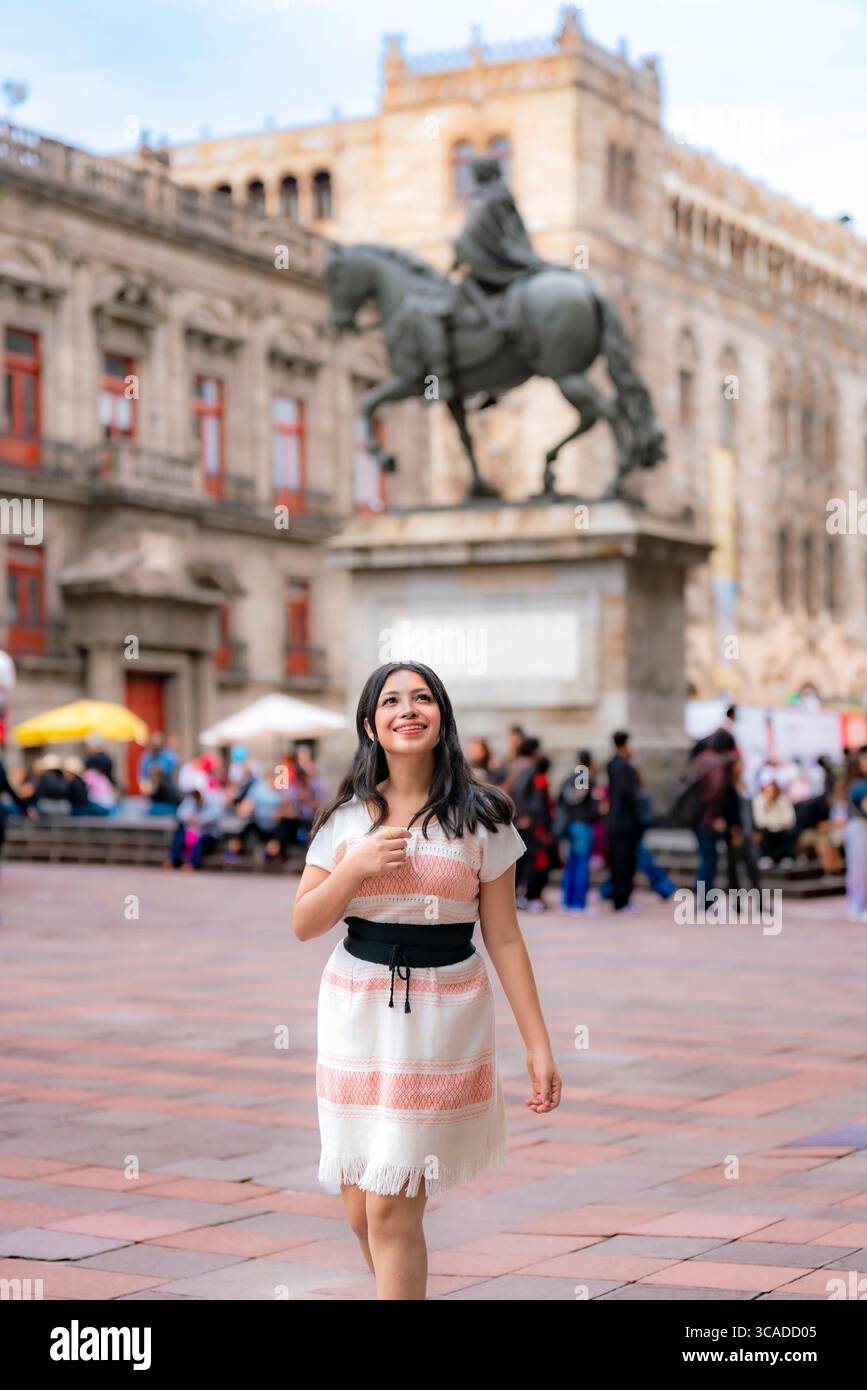 Young happy Latina woman with a bright smile walks confidently in front of the statue of Carlos IV, wearing a white and coral traditional dress in Mex Stock Photo