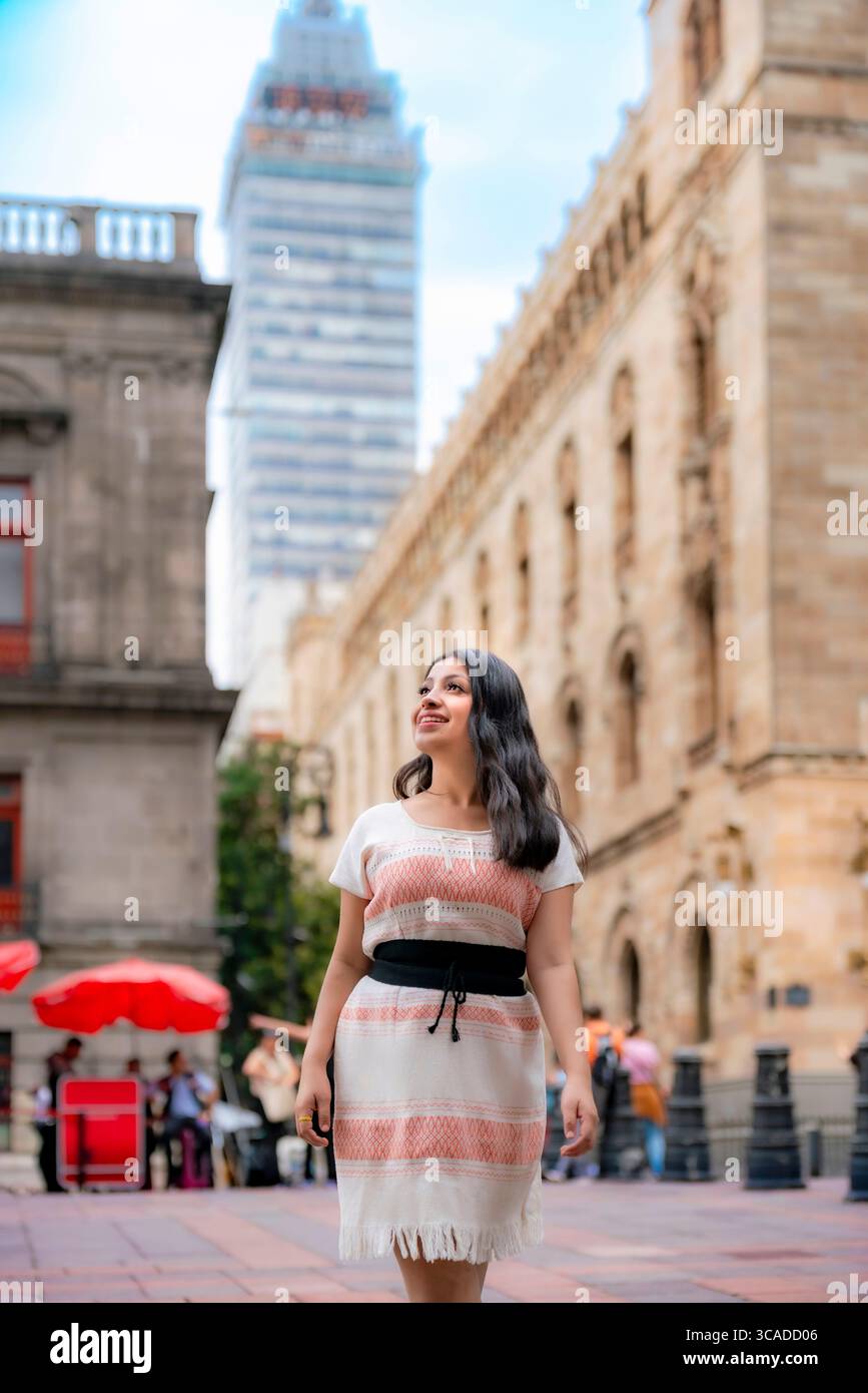 Happy young Latina woman in a white and coral woven dress with a black sash walks through downtown Mexico City near the Torre Latinoamericana, smiling Stock Photo