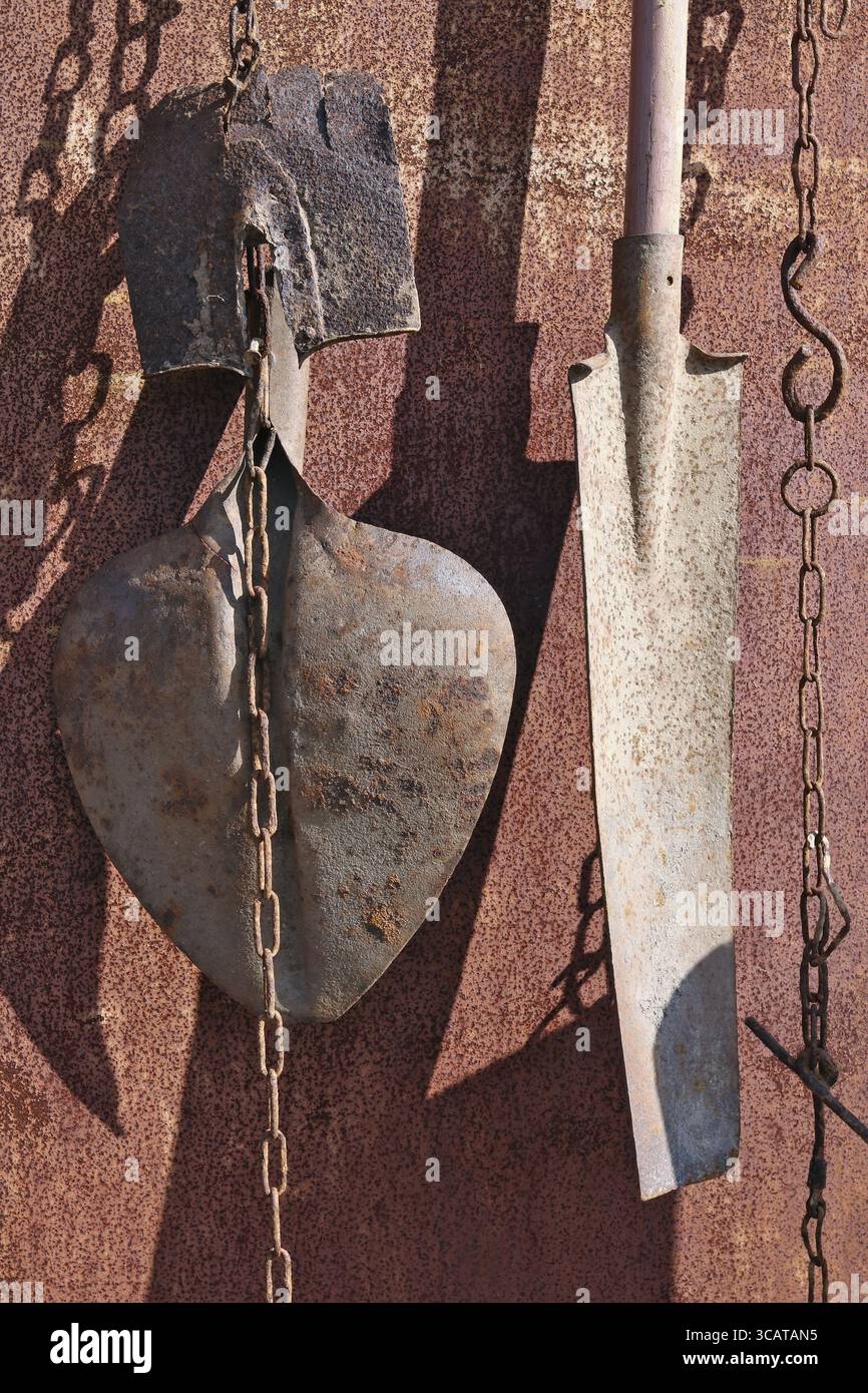 On the rusty metal wall of the village shed hang on the chains used old vintage agricultural shovels. Sunny spring day shot Stock Photo