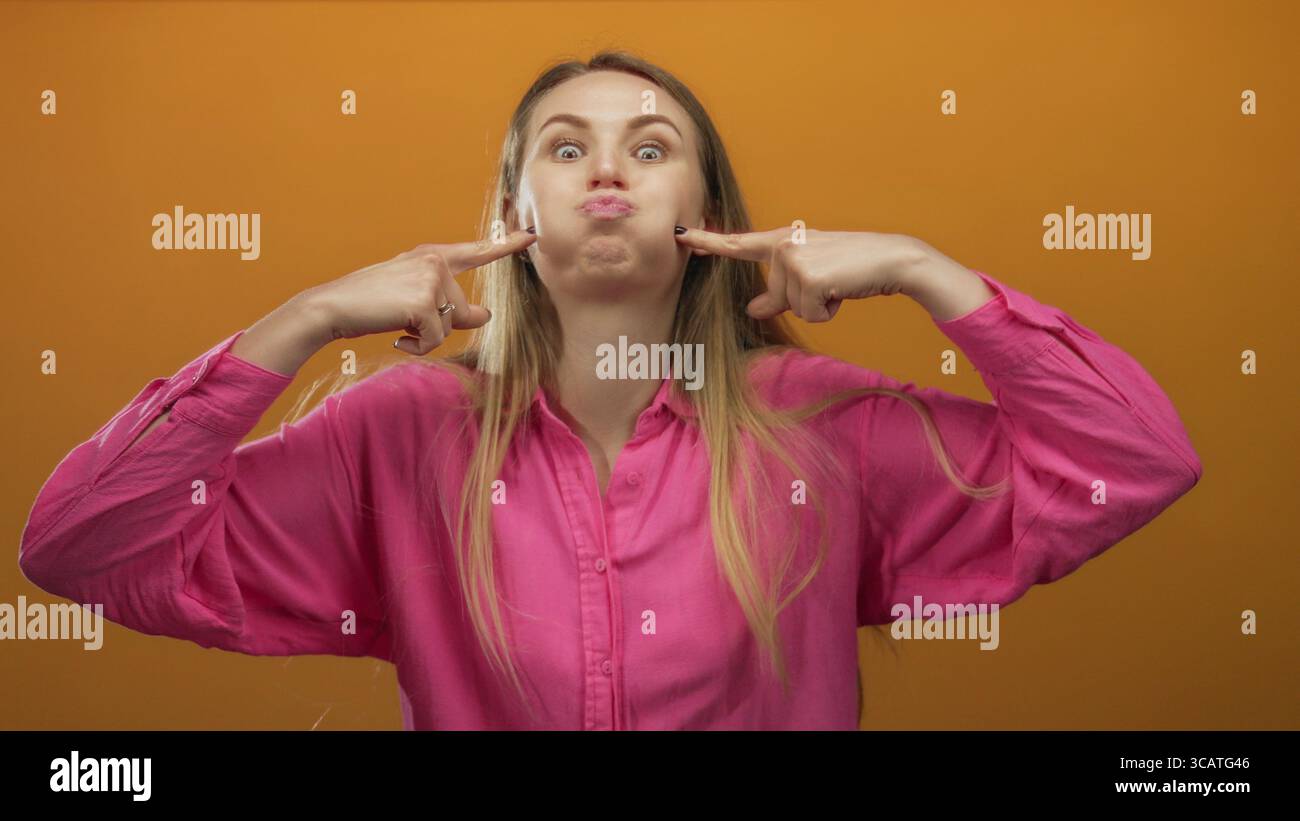 Woman with long hair and pink shirt playfully puffing cheeks against an isolated orange background, expressing silly emotions with a joyful expression Stock Photo