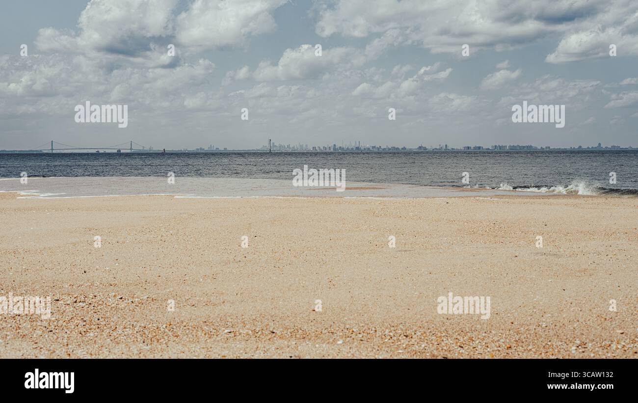 Distant view of the Manhattan skyline across a quiet beach and calm ocean, with the Verrazzano-Narrows Bridge visible from Sandy Hook, New Jersey. Stock Photo