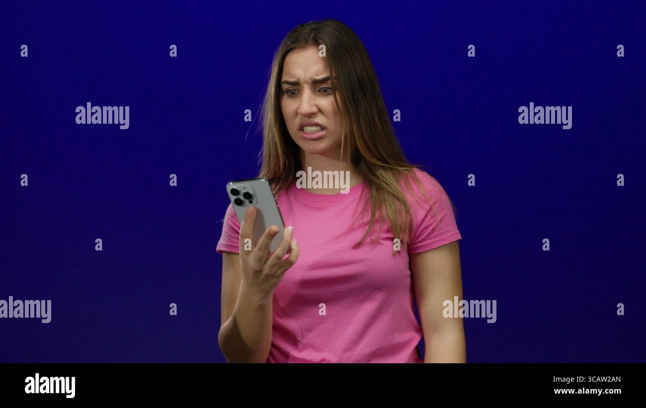 Woman expressing frustration while holding smartphone against blue background, wearing pink shirt, showcasing emotions of annoyance and confusion in a Stock Photo
