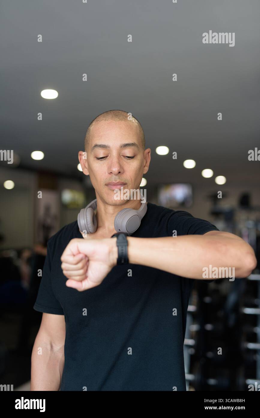 Hispanic bald man athlete in gym using smartwatch fitness tracker monitoring his exercise stats and heart rate with wearable technology in modern gym Stock Photo