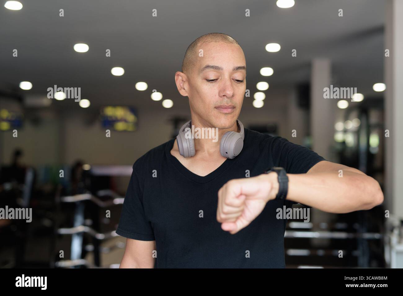 Hispanic bald man athlete in gym using smartwatch fitness tracker monitoring his exercise stats and heart rate with wearable technology in modern gym Stock Photo