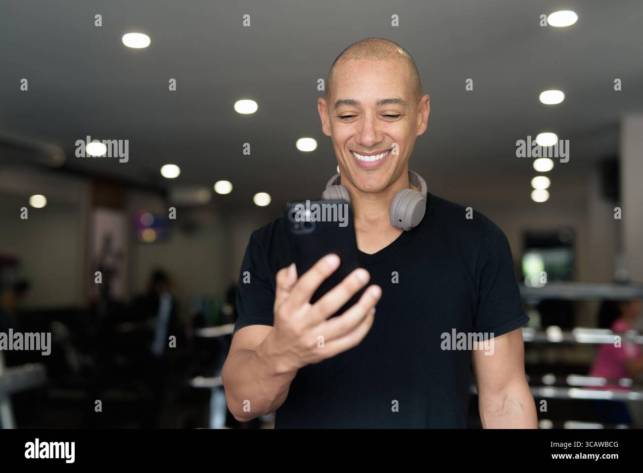 Bald Hispanic man training in gym for fitness and workout. Muscular male exercising indoors with confidence, wearing black shirt and headphones. Healt Stock Photo