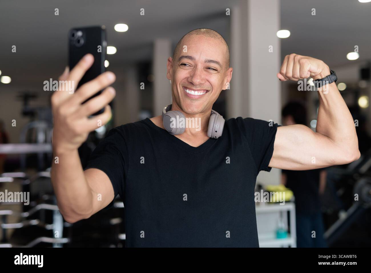 Bald Hispanic man training in gym for fitness and workout. Muscular male exercising indoors with confidence, wearing black shirt and headphones. Healt Stock Photo