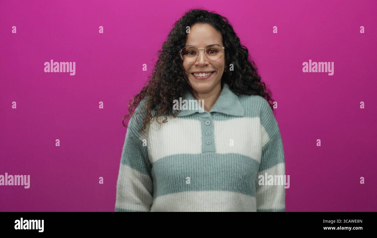 Woman smiling confidently in glasses against vibrant pink background expressing varied emotions with curly hair and warm sweater showcasing casual sty Stock Photo