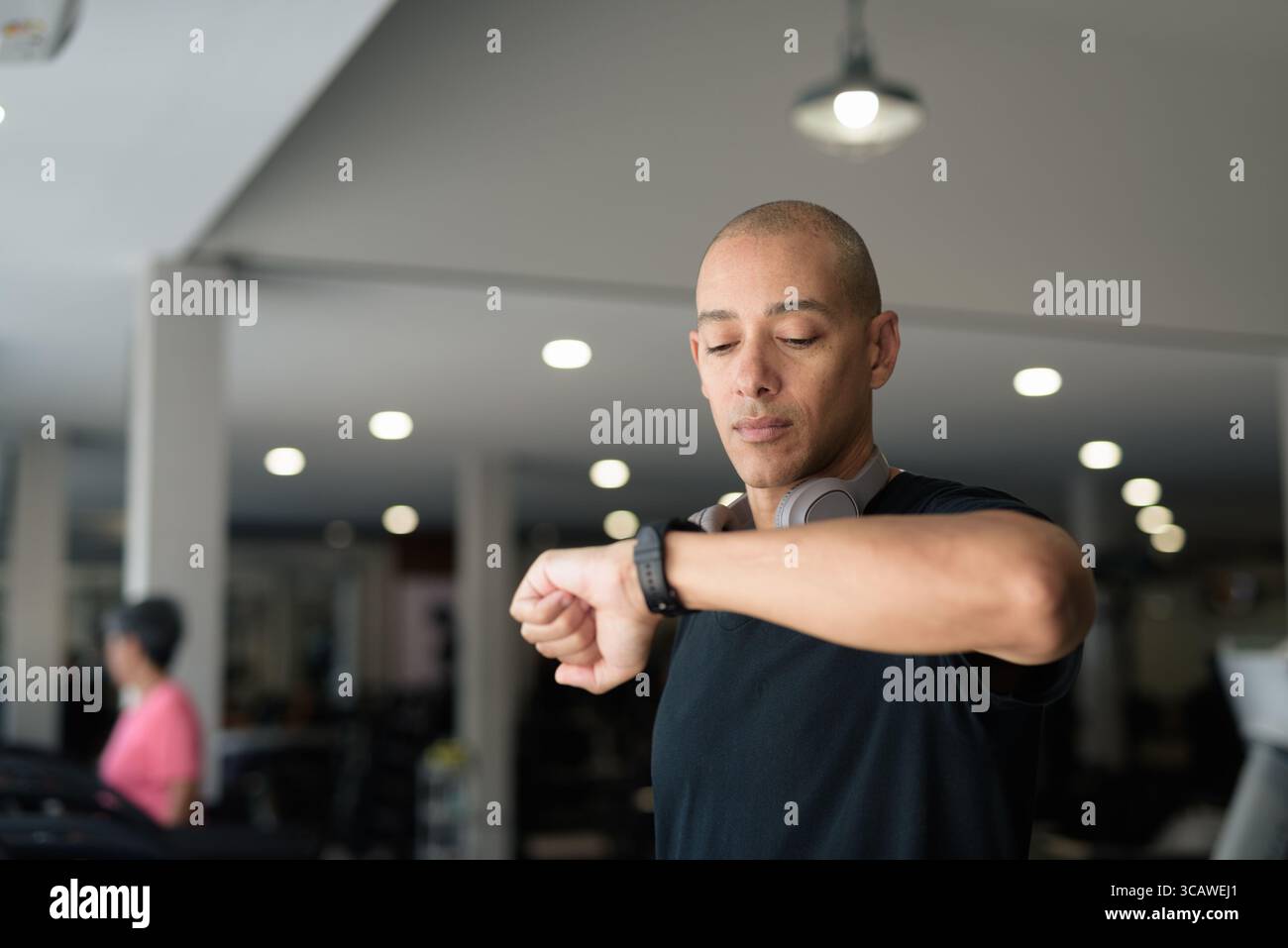 Hispanic bald man athlete in gym using smartwatch fitness tracker monitoring his exercise stats and heart rate with wearable technology in modern gym Stock Photo