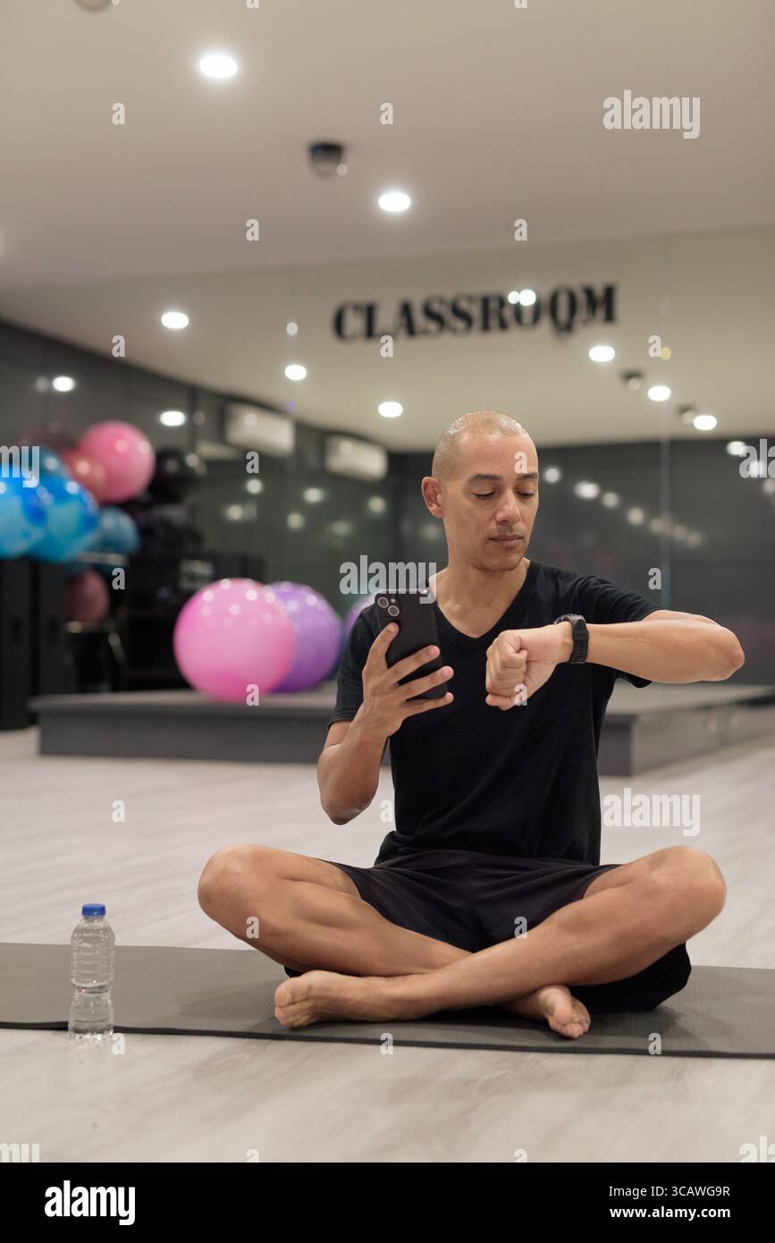 Hispanic bald man athlete in gym using smartwatch fitness tracker monitoring his exercise stats and heart rate with wearable technology in modern gym Stock Photo