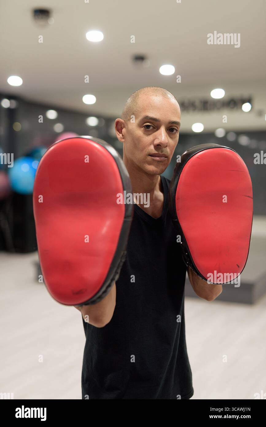 Hispanic bald male fitness trainer wearing black shirt holding Muay Thai kick pads in modern gym. Martial arts, combat sports, striking training, and Stock Photo