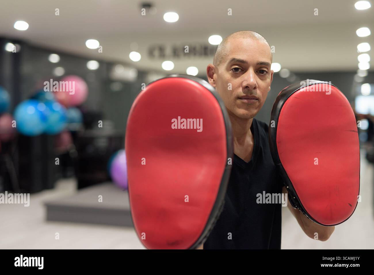 Hispanic bald male fitness trainer wearing black shirt holding Muay Thai kick pads in modern gym. Martial arts, combat sports, striking training, and Stock Photo