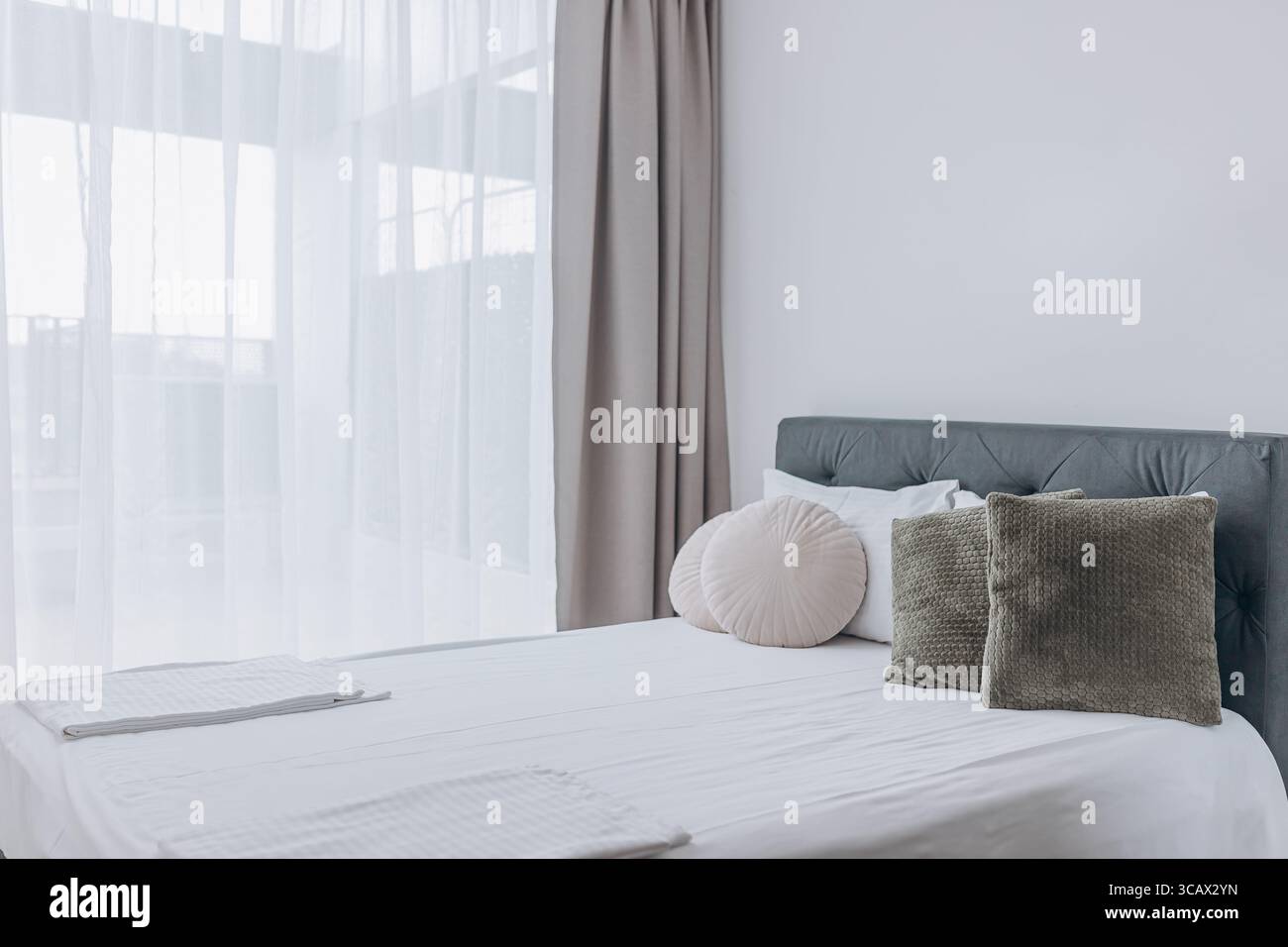 Bright bedroom with large window, beige curtains, and round decorative pillows on white bed. Stock Photo