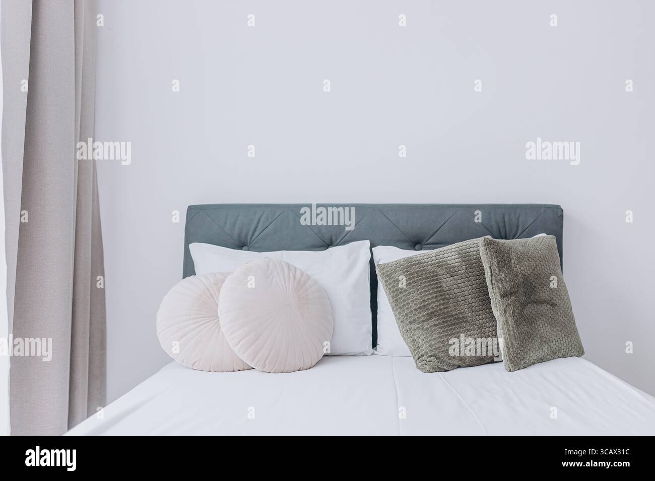 Symmetrical arrangement of decorative pillows on soft white bed with fabric headboard and curtains. Stock Photo