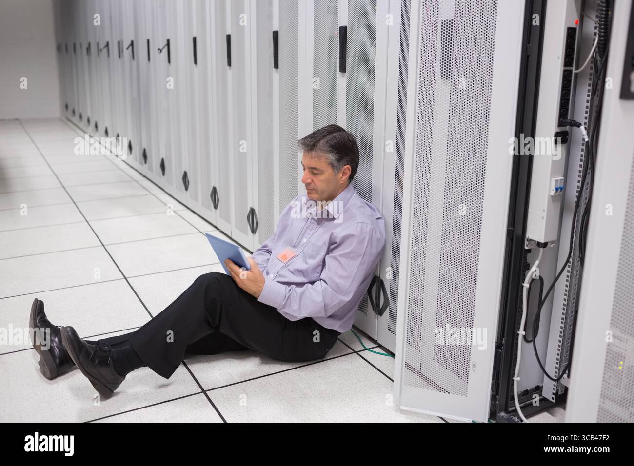 Middle-aged man IT technician sitting beside server rack in server room using tablet, copy space Stock Photo