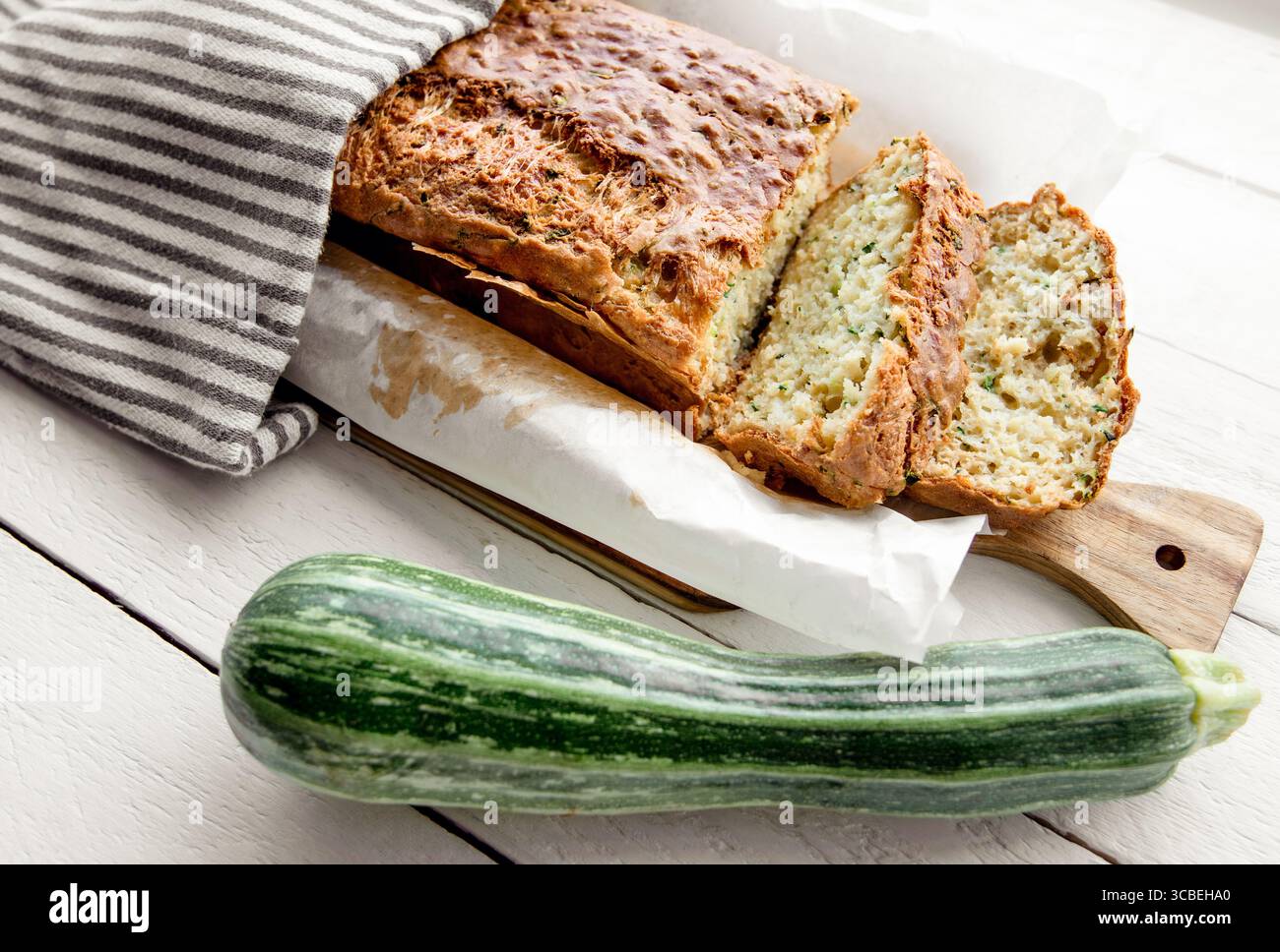 Zucchini and cheese bread recipe, a loaf of bread on a wooden cutting board, covered with a kitchen towel, with a whole zucchini vegetable next to it. Stock Photo